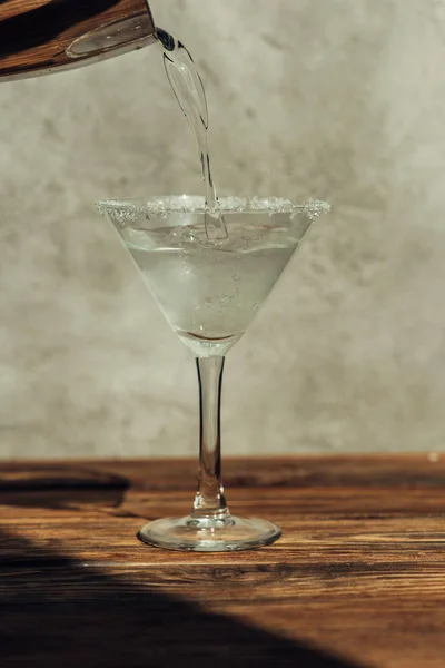 Liquid pouring from shaker into martini glass decorated with sugar on wooden surface in sunlight — Stock Photo