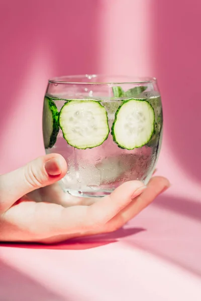 Cropped view of woman holding glass of water with cucumber slices in sunlight on pink background — Stock Photo