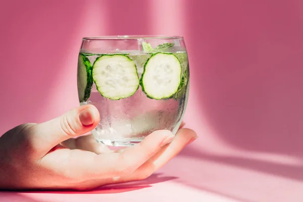 Vue recadrée d'une femme tenant un verre d'eau avec des tranches de concombre au soleil sur fond rose — Photo de stock