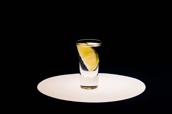 Tequila fresco con lima sobre círculo iluminado aislado sobre negro - foto de stock