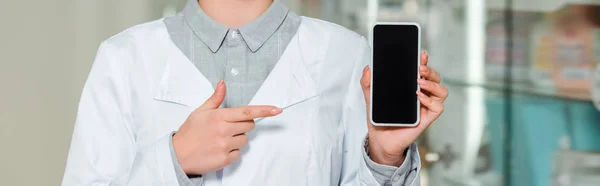 Vue recadrée du pharmacien pointant du doigt le smartphone avec écran blanc, prise de vue panoramique — Photo de stock