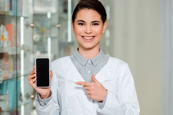 Улыбающийся фармацевт указывает пальцем на смартфон с пустым экраном — стоковое фото
