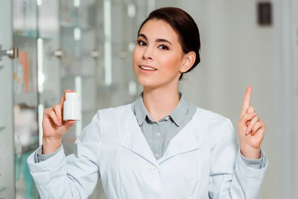 Schöne Apothekerin zeigt mit dem Finger, während sie Glas mit Pillen in der Hand hält und in die Kamera schaut — Stockfoto