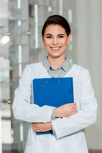 Attraktive Apothekerin mit Klemmbrett lächelt in Apotheke in die Kamera — Stockfoto