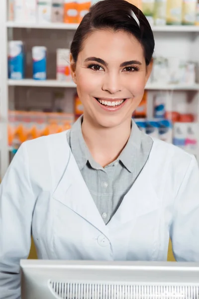 Farmacêutico atraente sorrindo para a câmera com medicamentos em prateleiras no fundo — Fotografia de Stock