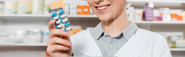 Vista recortada del farmacéutico sonriente sosteniendo pastillas en la farmacia, tiro panorámico - foto de stock