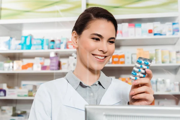 Belo farmacêutico sorridente segurando blister com pílulas na farmácia — Fotografia de Stock