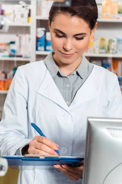 Attractive pharmacist writing on clipboard at pharmacy counter — Stock Photo