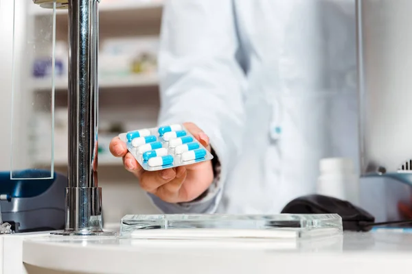 Обрезанный вид аптекаря, держащего волдырь с таблетками у аптекарского прилавка — стоковое фото