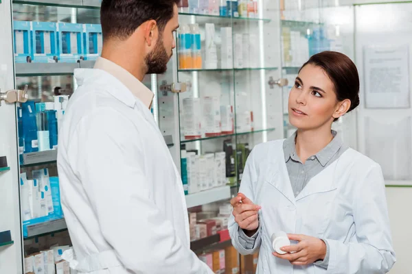 Pharmacist with jar of pills looking at colleague in pharmacy — Stock Photo