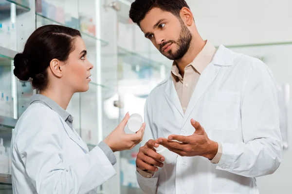 Pharmacist showing to colleague jar of pills by pharmacy showcase — Stock Photo