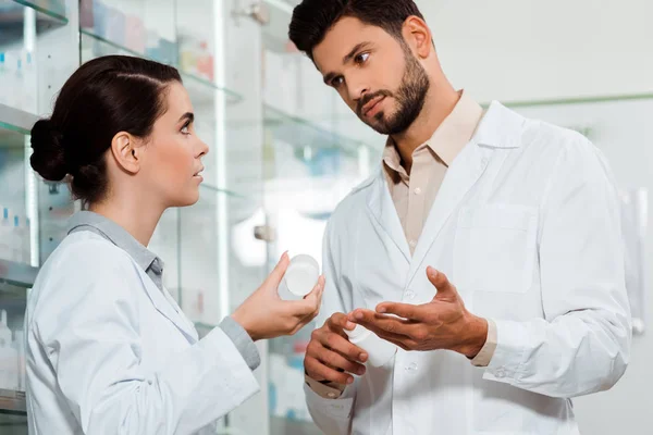 Pharmacist showing to colleague jar of pills by pharmacy showcase — Stock Photo