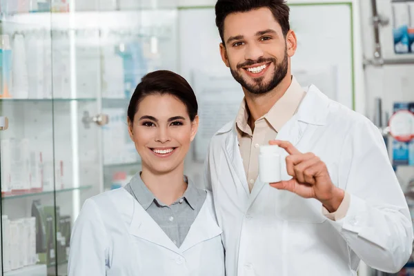 Farmacéuticos con frasco de pastillas sonriendo a cámara en farmacia — Stock Photo