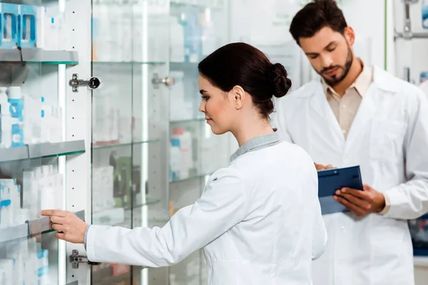 Pharmacien prenant des médicaments de vitrine à côté de son collègue avec presse-papiers en pharmacie — Photo de stock