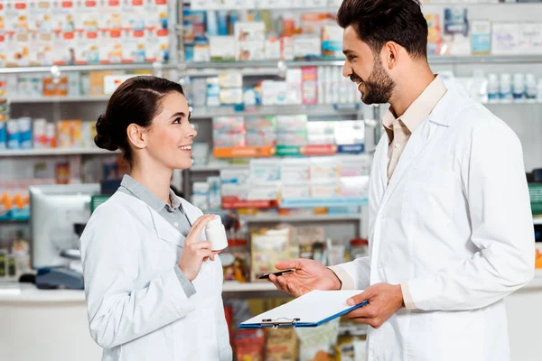 Vista lateral de farmacêuticos com pílulas e prancheta sorrindo uns para os outros na farmácia — Fotografia de Stock