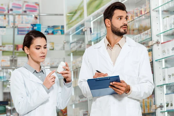 Farmacêutico com prancheta e colega com pílulas por vitrine farmácia — Fotografia de Stock