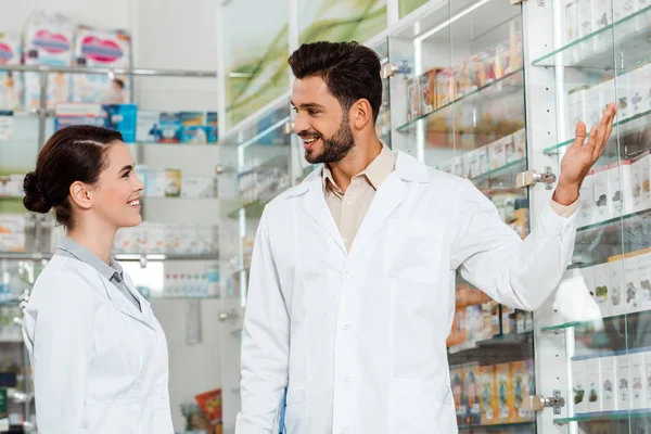 Side view of handsome pharmacist pointing at showcase to colleague in drugstore — Stock Photo