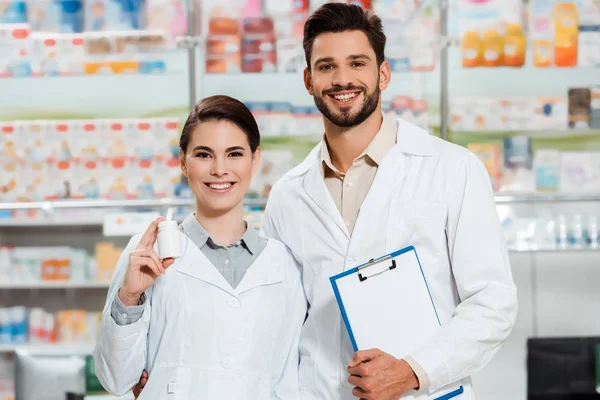 Lächelnder Apotheker mit Klemmbrett und Pillen in der Apotheke vor laufender Kamera — Stockfoto