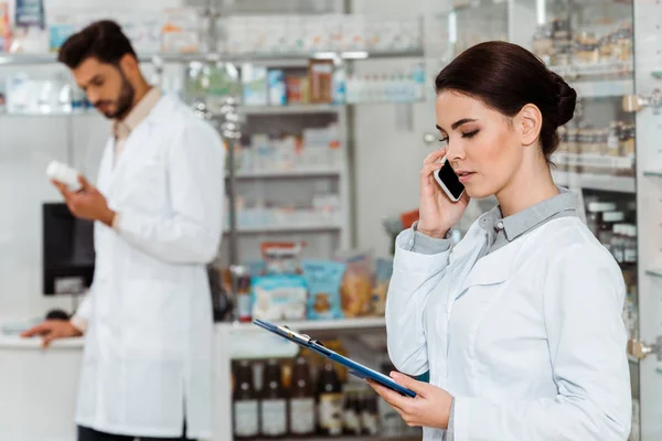 Foco seletivo do farmacêutico com área de transferência falando no smartphone com colega em segundo plano — Fotografia de Stock