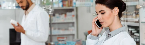 Panoramic shot of pharmacist talking on smartphone with colleague at background — Stock Photo