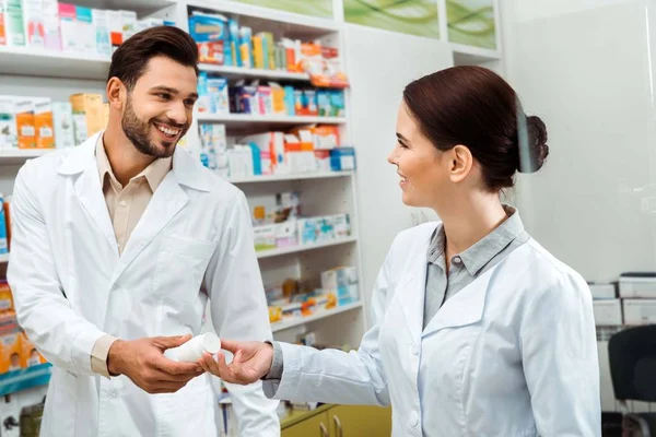 Farmacéutico sonriente dando a colega frasco con pastillas en la farmacia - foto de stock