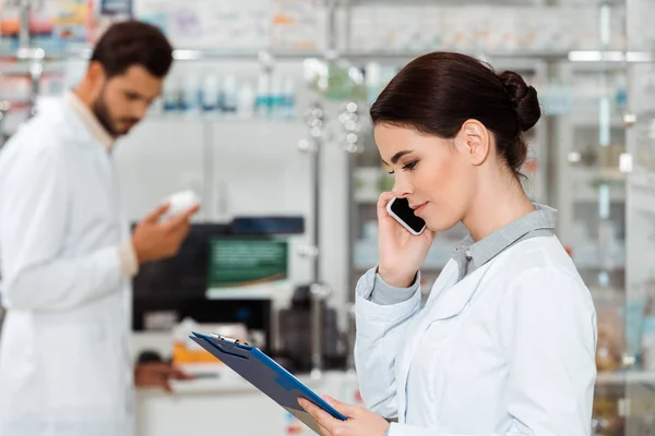 Focus selettivo del farmacista con appunti che parla su smartphone con colleghi che tengono pillole in background — Foto stock