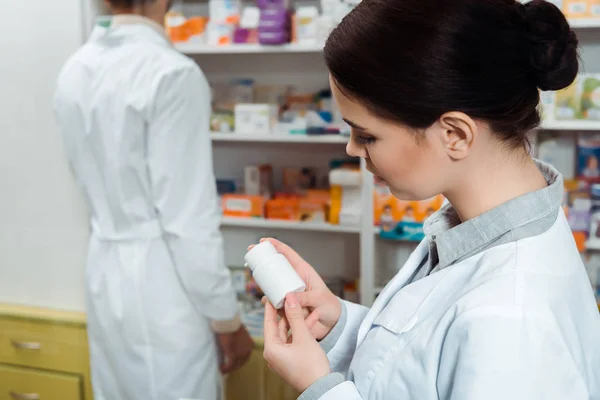 Foco seletivo do farmacêutico olhando para frasco de pílulas com colega em segundo plano — Fotografia de Stock