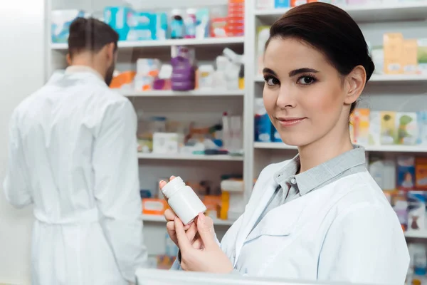 Farmacêutico segurando frasco de pílulas enquanto olha para a câmera com colega no fundo — Fotografia de Stock