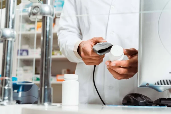 Vista recortada de las píldoras de escaneo del farmacéutico con el escáner de código de barras en la farmacia - foto de stock