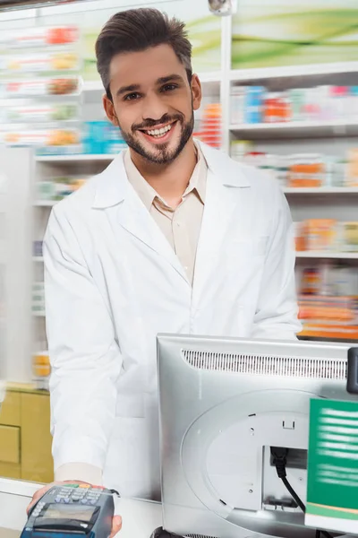Guapo farmacéutico sonriente sosteniendo terminal en el mostrador de la farmacia - foto de stock