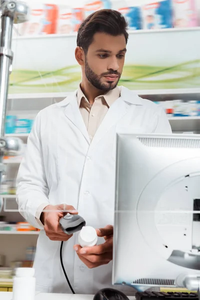 Красивый фармацевт с таблетками и сканером штрих-кодов смотрит на компьютер в аптеке — стоковое фото