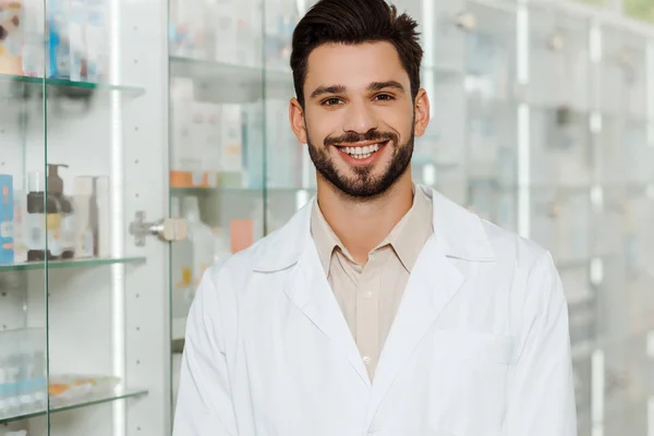 Handsome druggist smiling at camera by pharmacy showcase — Stock Photo