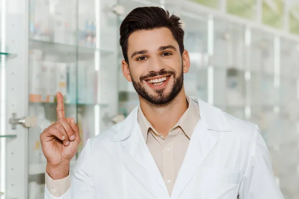 Guapo farmacéutico sonriendo a la cámara y señalando con el dedo en la farmacia - foto de stock