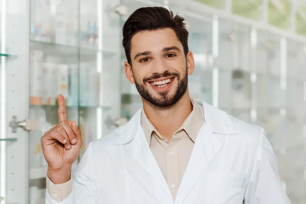 Bonito farmacêutico sorrindo para a câmera e apontando com o dedo na farmácia — Fotografia de Stock