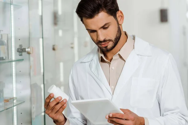 Farmacêutico bonito usando comprimido digital enquanto segurando frasco de pílulas — Fotografia de Stock