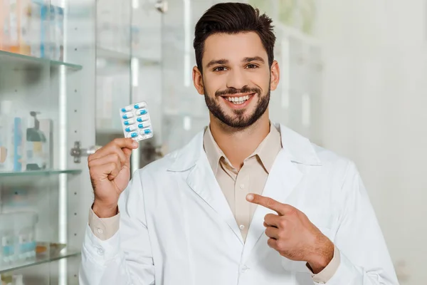 Handsome pharmacist smiling at camera and pointing at blister with pills — Stock Photo
