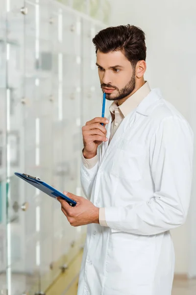 Pharmacien pensif avec presse-papiers et stylo en pharmacie — Photo de stock