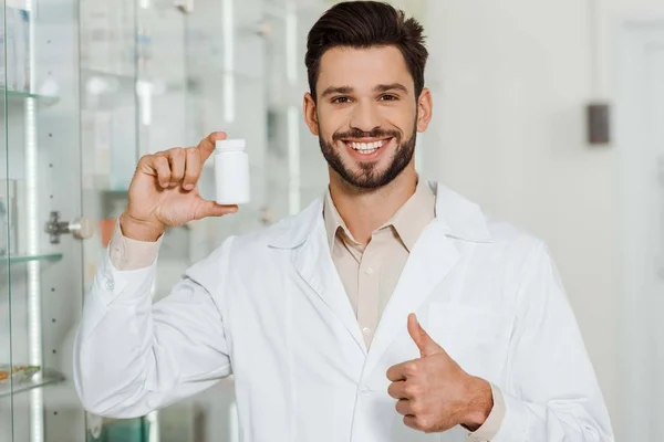 Фармацевт с банкой таблеток улыбается в камеру и показывает большой палец вверх — стоковое фото