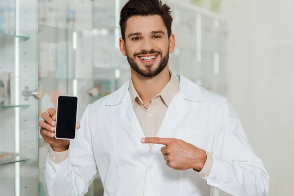 Улыбающийся фармацевт указывает на смартфон с пустым экраном в аптеке — стоковое фото