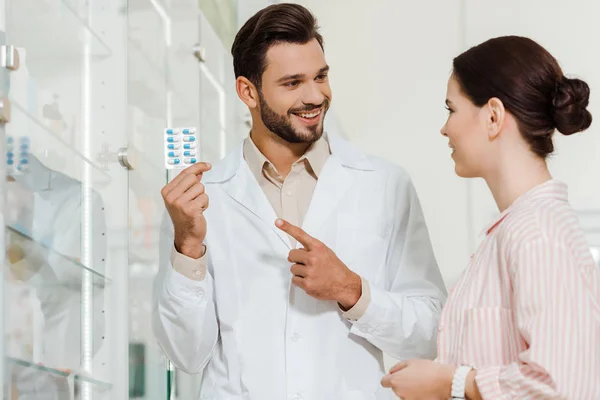 Drogadicto sonriente señalando ampollas con pastillas al cliente en la farmacia - foto de stock