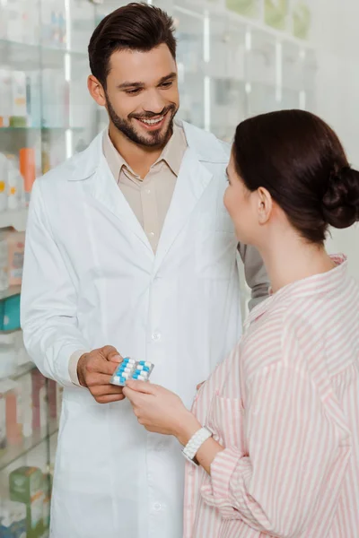 Droguiste souriant donnant blister avec des pilules au client par vitrine de pharmacie — Photo de stock