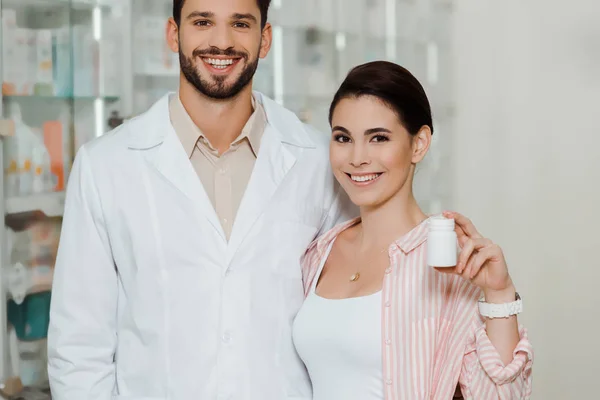 Cliente atractivo mostrando frasco con pastillas al lado del farmacéutico sonriente - foto de stock