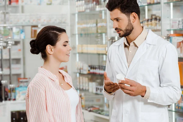 Vue latérale du pharmacien montrant au client pot de pilules par vitrine de pharmacie — Photo de stock