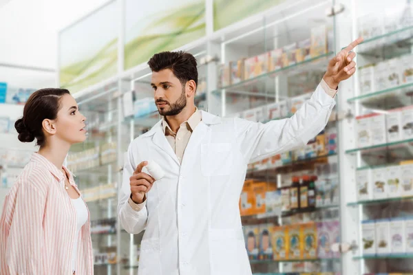 Handsome pharmacist pointing at showcase to customer — Stock Photo