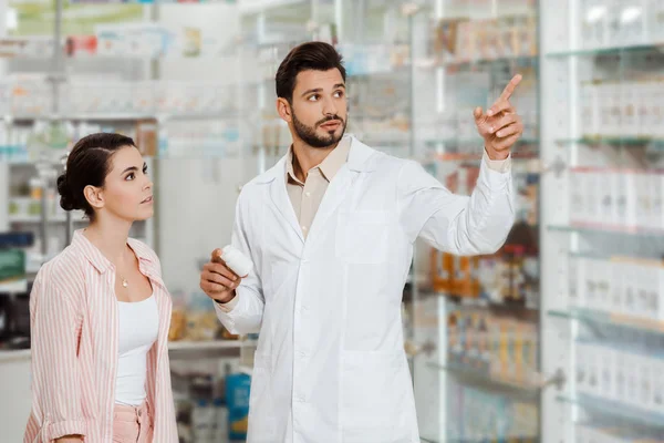 Druggist with jar of pills pointing at pharmacy showcase to customer — Stock Photo