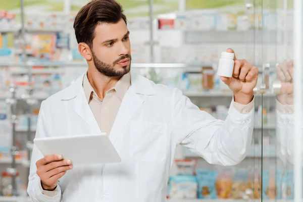Handsome druggist holding digital tablet and looking at jar of pills — Stock Photo