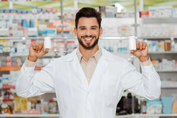 Handsome pharmacist smiling at camera and holding jars with pills — Stock Photo