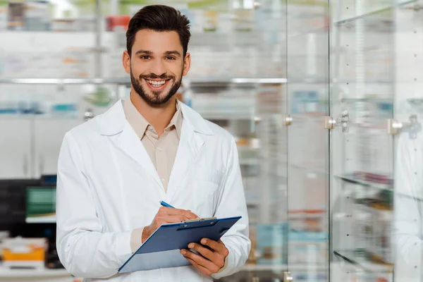 Handsome pharmacist with clipboard smiling at camera by drugstore showcase — Stock Photo