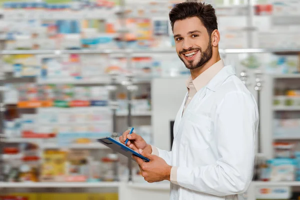 Selektiver Fokus eines lächelnden Apothekers mit Klemmbrett, der in der Apotheke in die Kamera blickt — Stockfoto