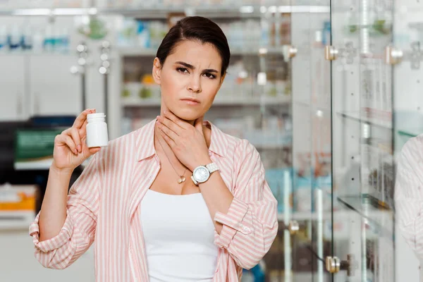 Woman with sore throat holding jar with pills with pharmacy showcase at background — Stock Photo